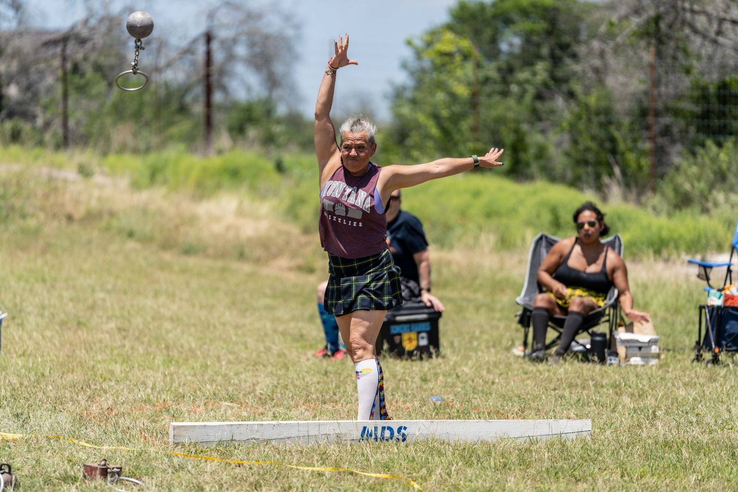 Athlete in kilt competes in Highland Games shot put, arm raised mid-throw on sunny grass field.