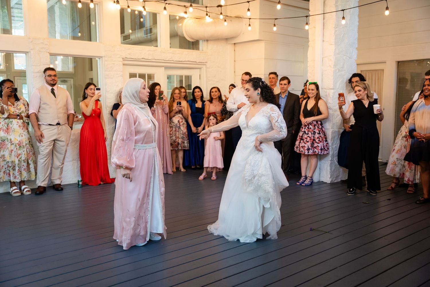 Bride in white gown dancing with guest in hijab at elegant indoor wedding reception with guests watching.