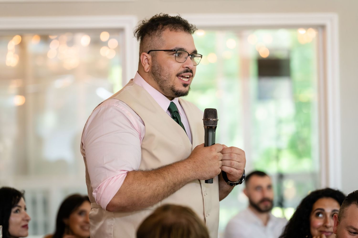 Man in pink shirt and tan vest speaks into microphone at elegant indoor wedding reception event.