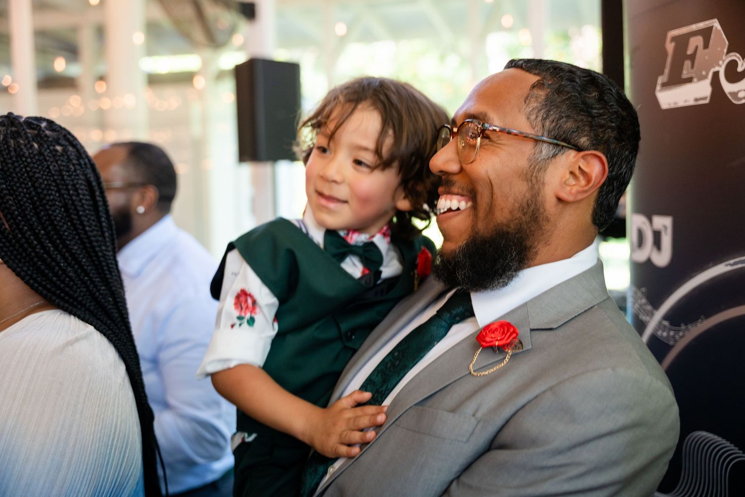 A man in a suit smiles while holding a young child in a green school uniform at an indoor event.