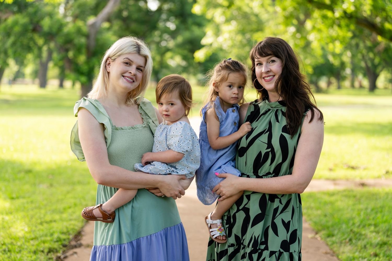 Two women holding young children smile outdoors on a park path surrounded by lush green trees.