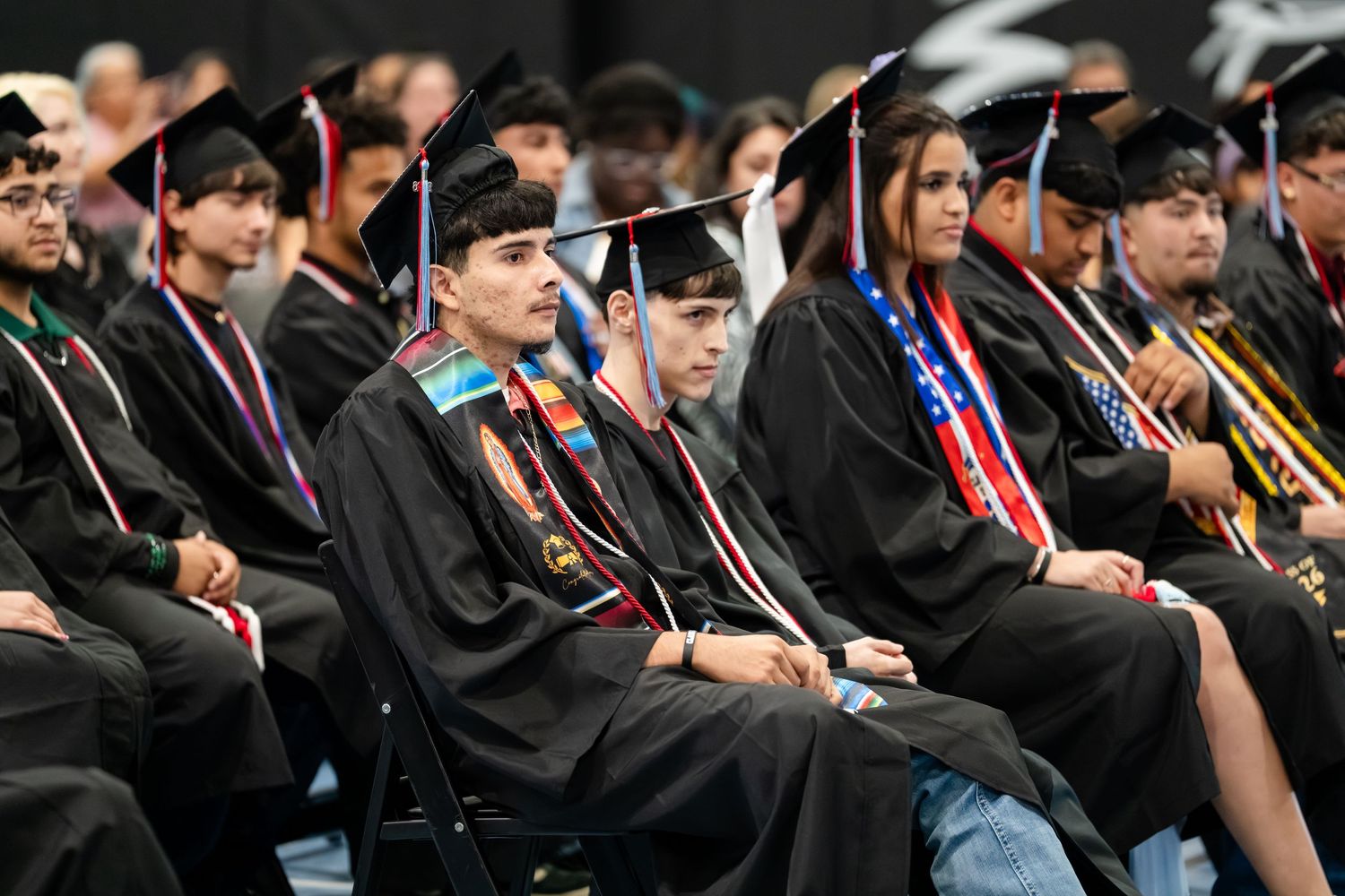 Students in black graduation gowns and caps seated at a commencement ceremony, some wearing colorful honor cords.