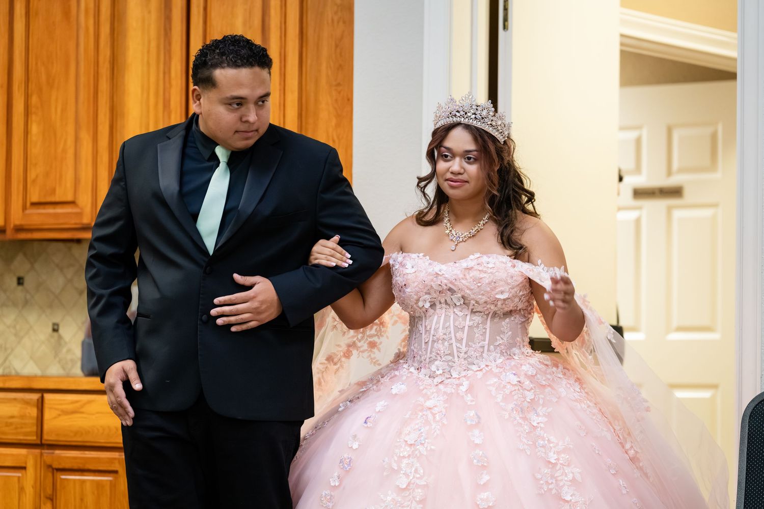 Young woman in pink quinceañera gown and tiara escorted by man in black suit at elegant celebration.