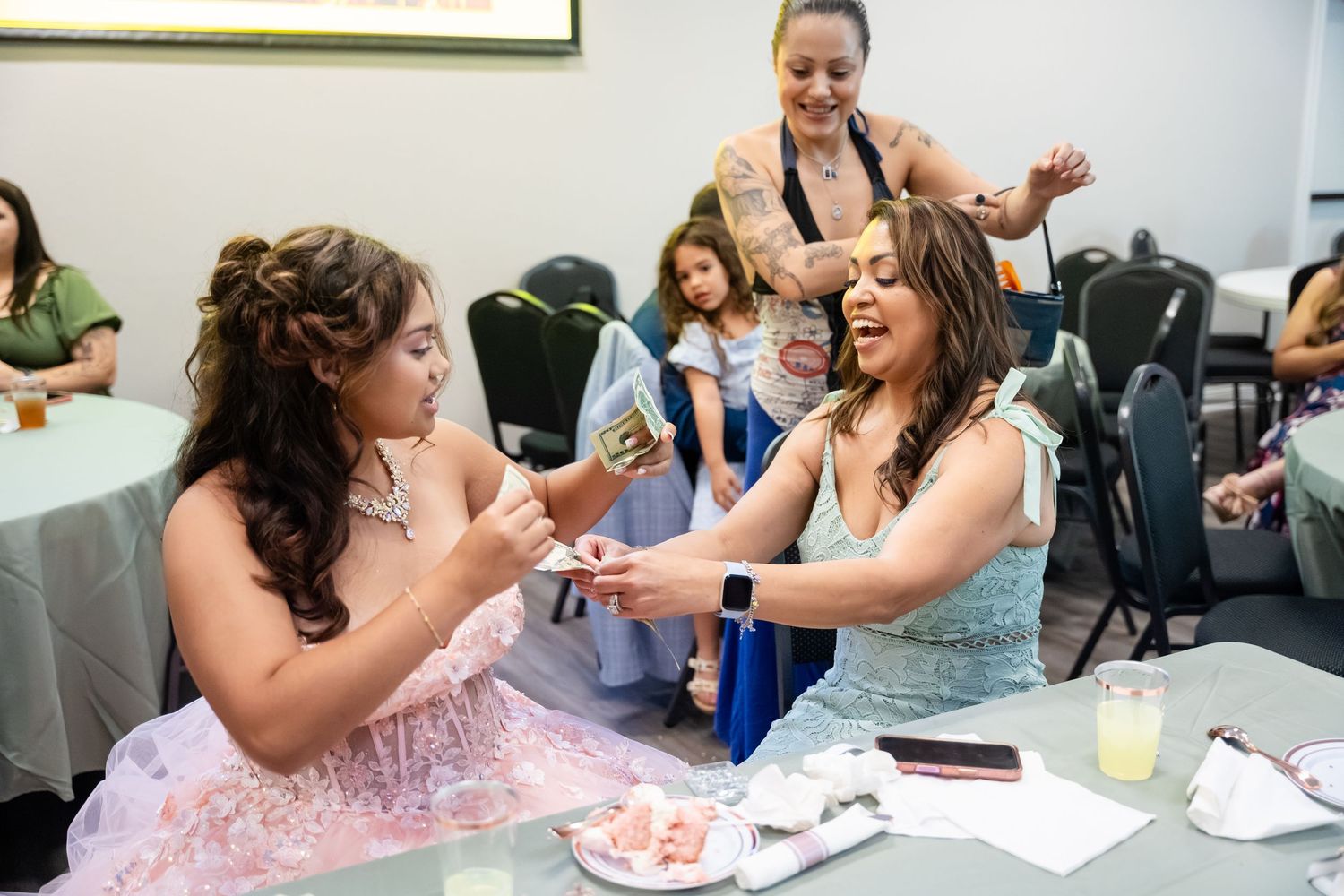 Two young women in formal dresses laugh and interact at a quinceañera celebration indoors.