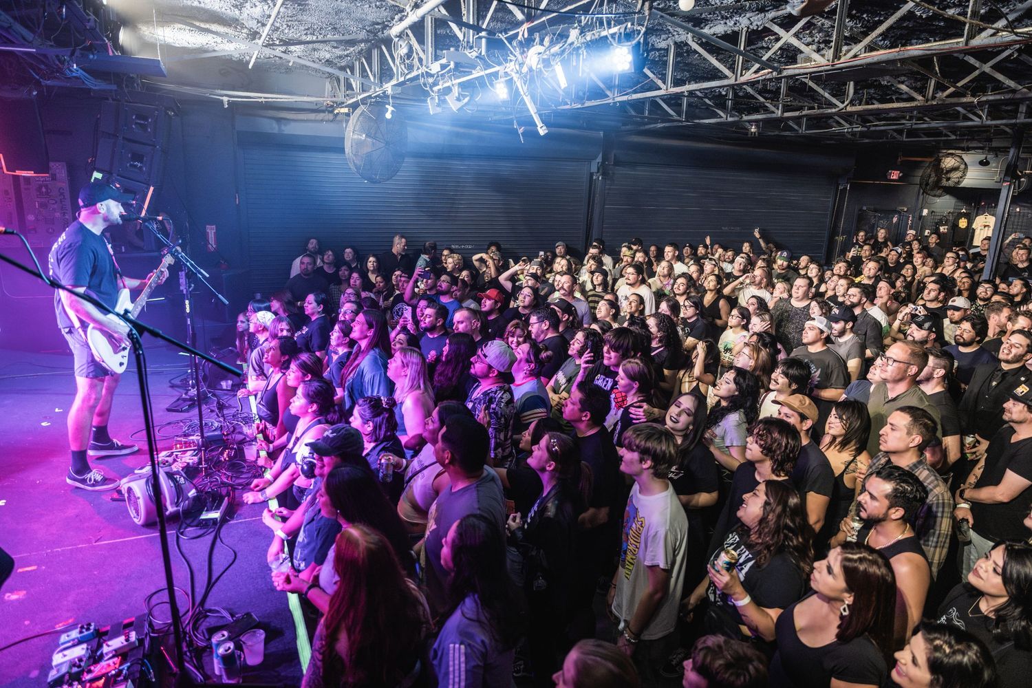 Concert crowd watching performers on stage with vibrant blue and purple lighting in a packed indoor venue.