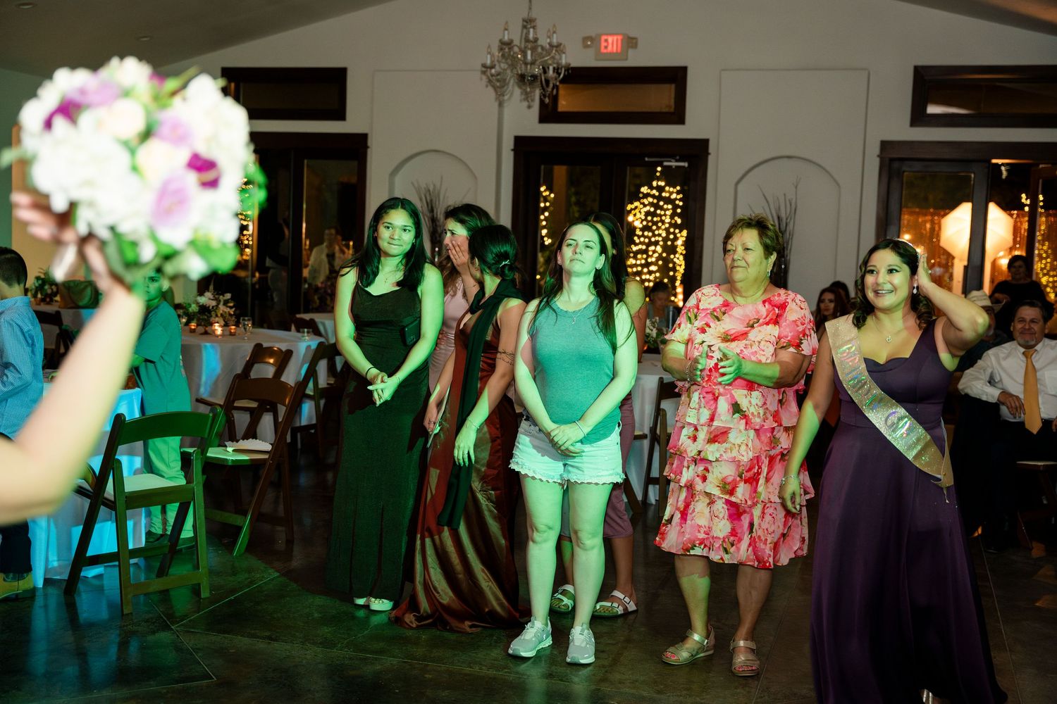 Wedding reception guests in green and floral dresses gather around for the bouquet toss at an evening event.