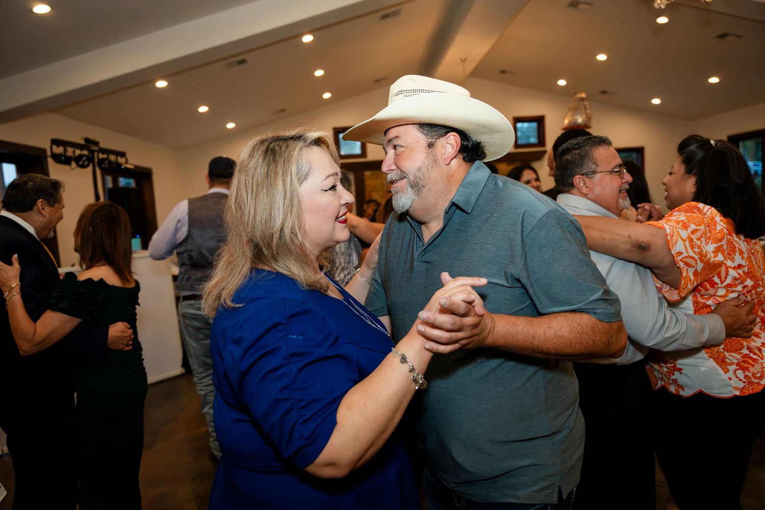 A couple dances together in a crowded indoor venue, with the man wearing a cowboy hat and teal shirt.