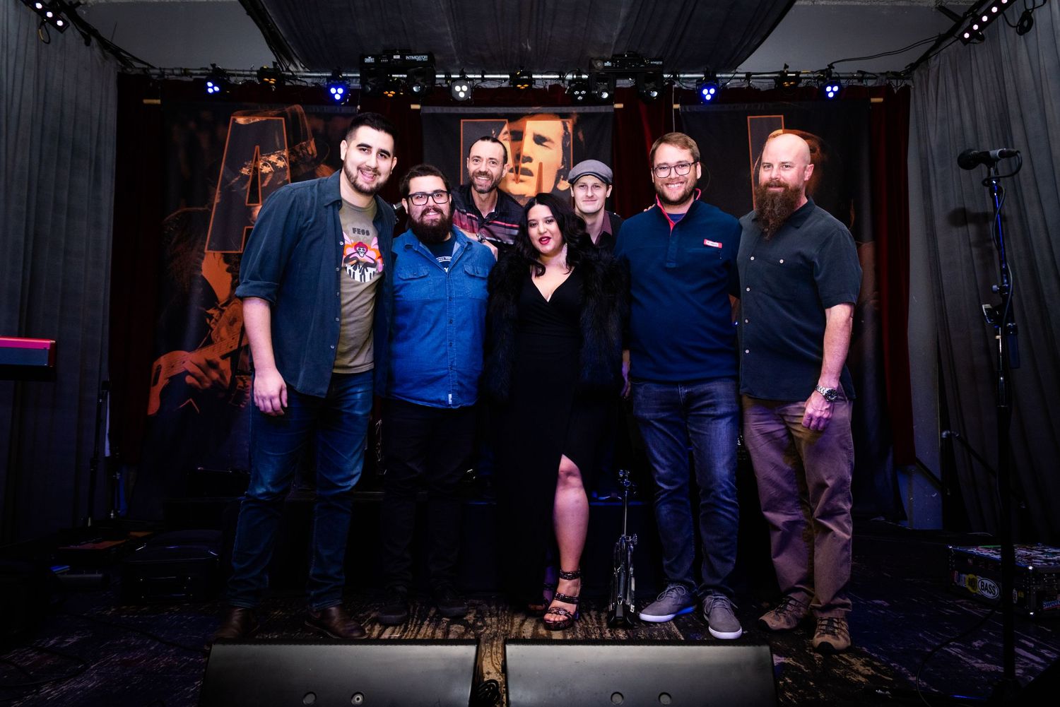 A group of musicians pose together on stage with instruments under dramatic lighting.