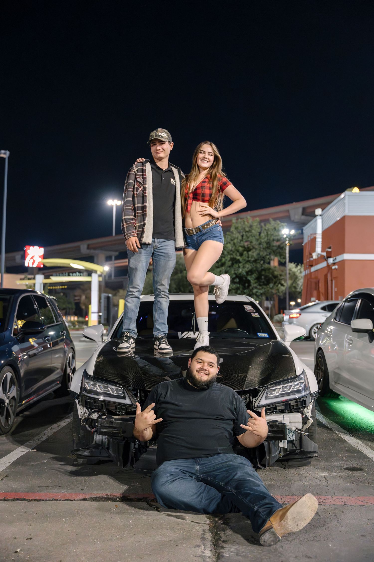 Three people pose on and around a black car at night in a parking lot.