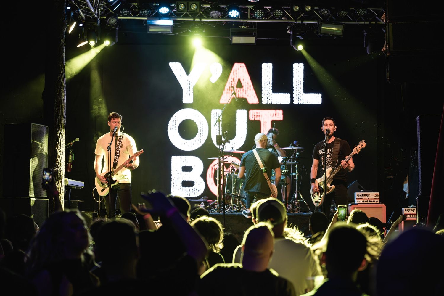 Band plays live show with 'Y'ALL OUT' sign glowing above silhouetted crowd.