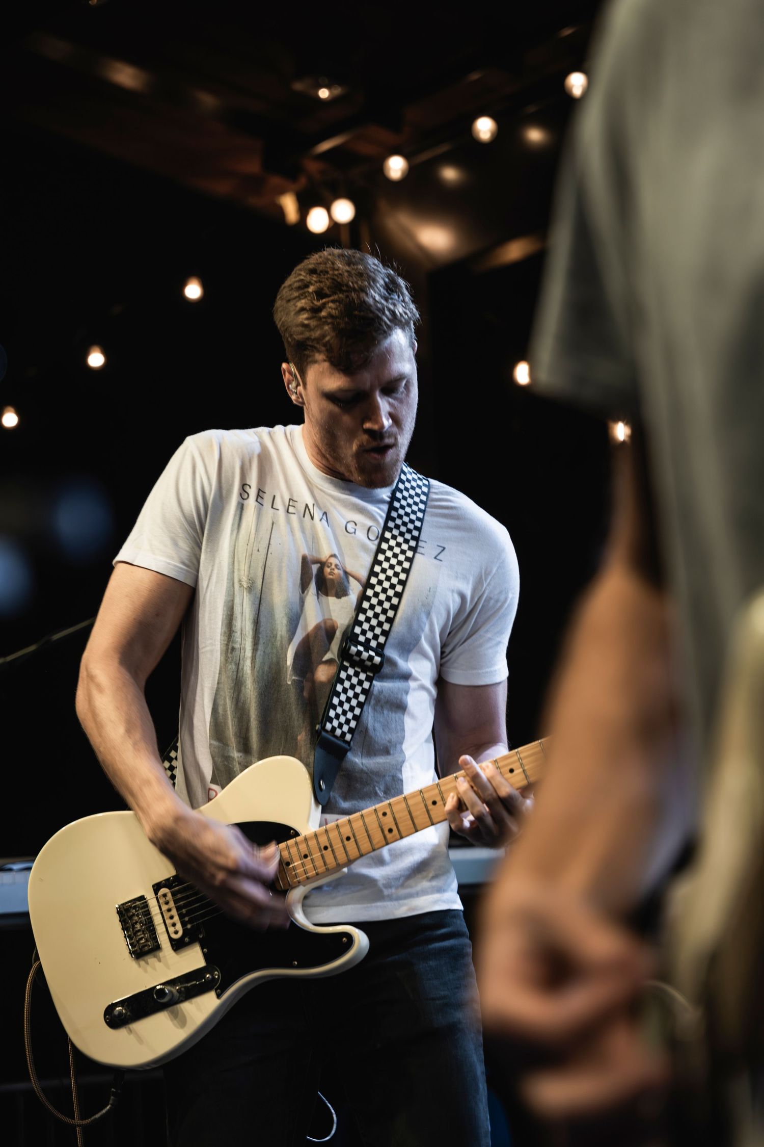 Guitarist in white t-shirt plays cream-colored Telecaster guitar under stage lighting.