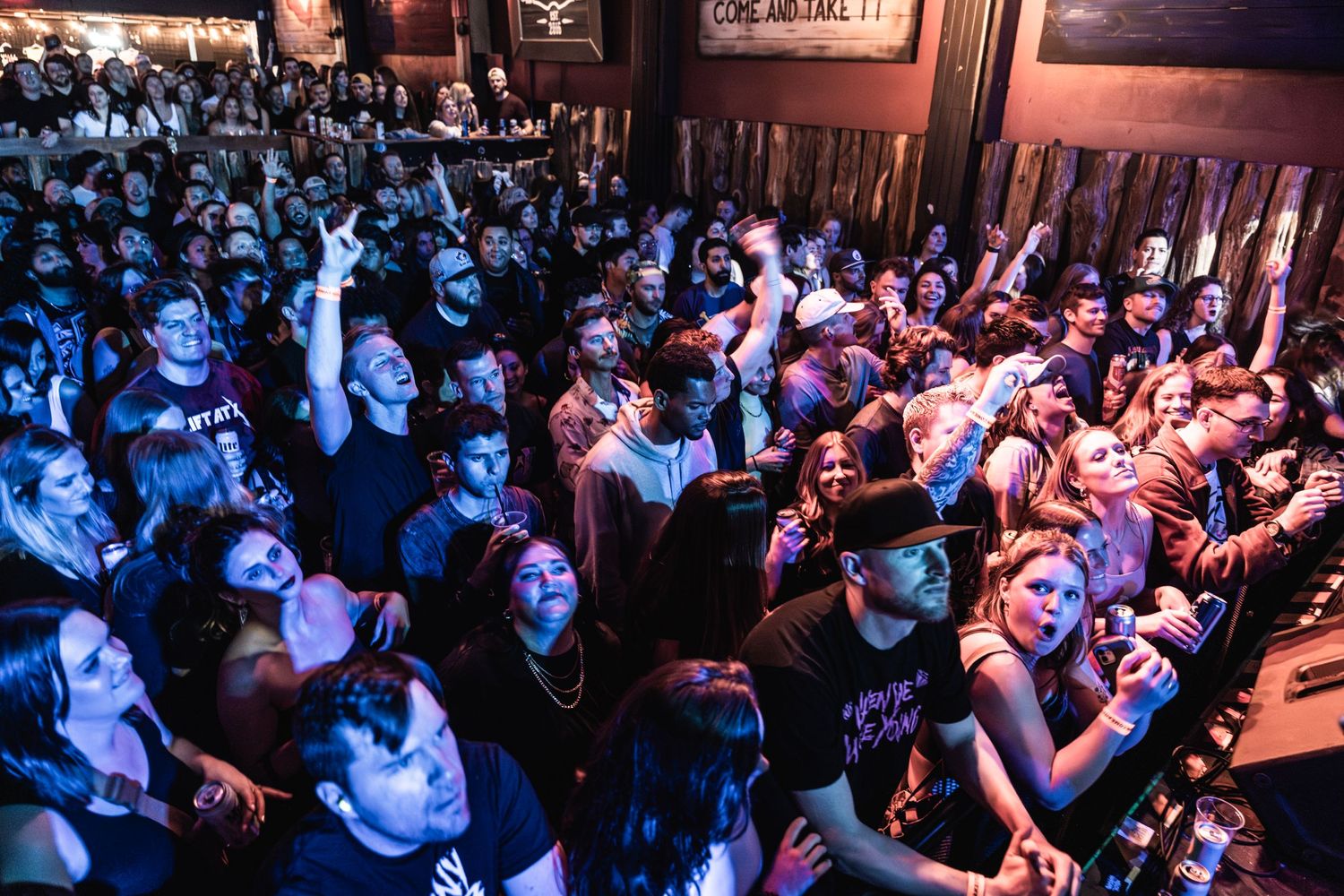 Large concert crowd with arms raised during performance in dimly lit venue.
