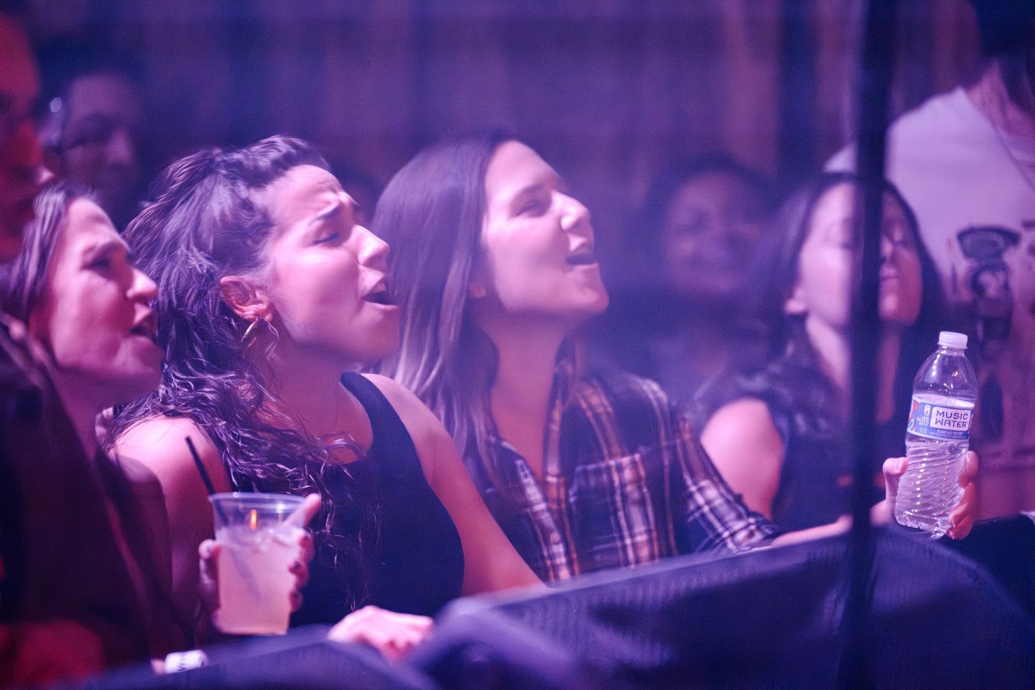 Concert attendees enjoying a performance with drinks in hand under purple venue lighting.