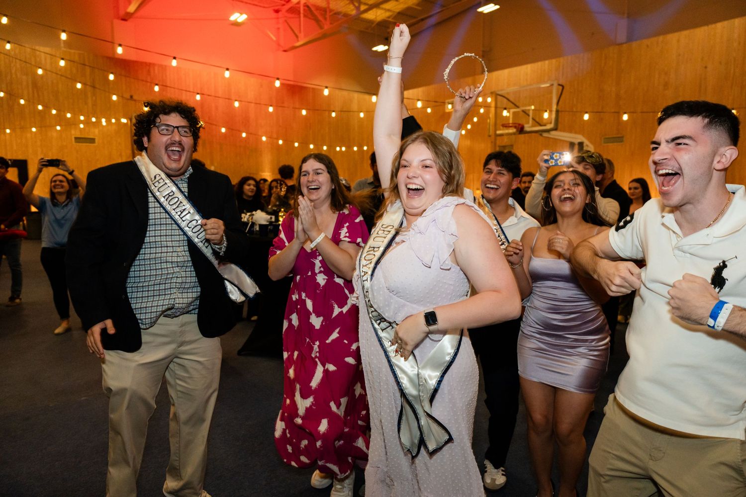A group dancing and celebrating at an indoor event with string lights and wooden walls.