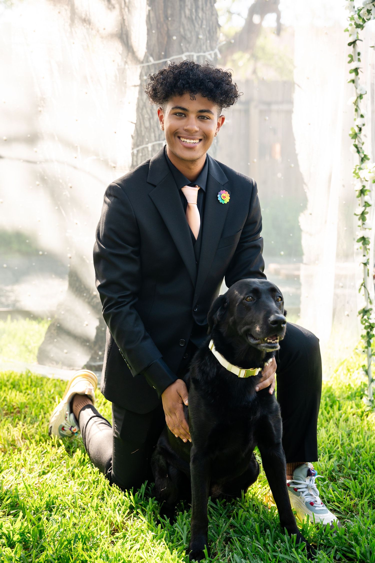 A person in formal attire kneels next to a black dog wearing a bright collar in a garden setting.