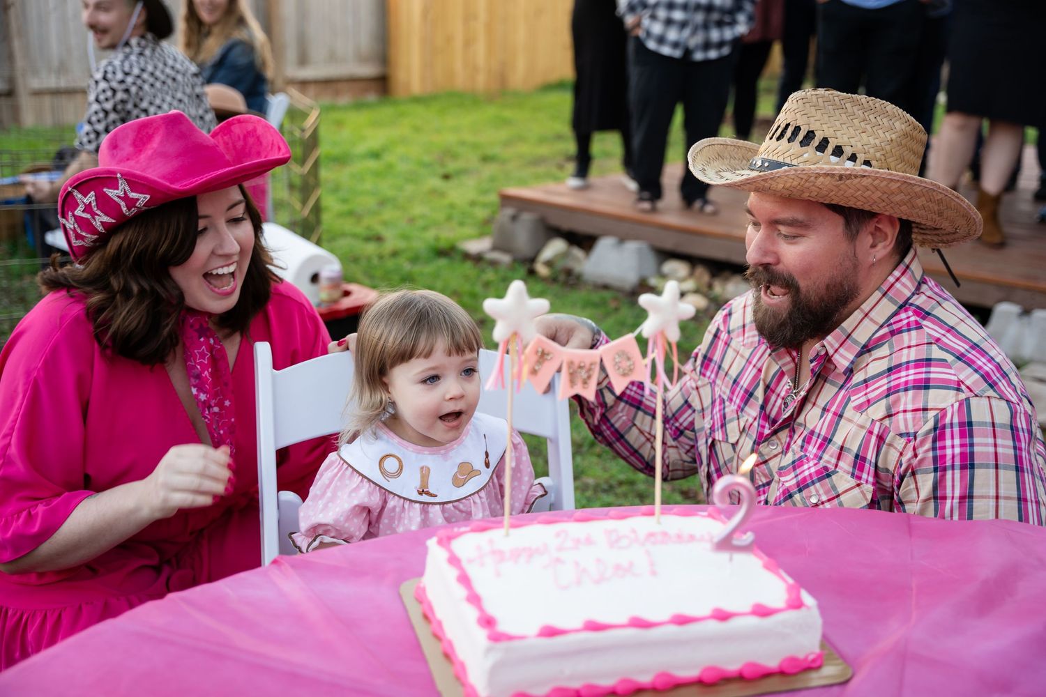 A pink birthday celebration in a backyard with a decorated cake on a table and smiling people in festive attire.