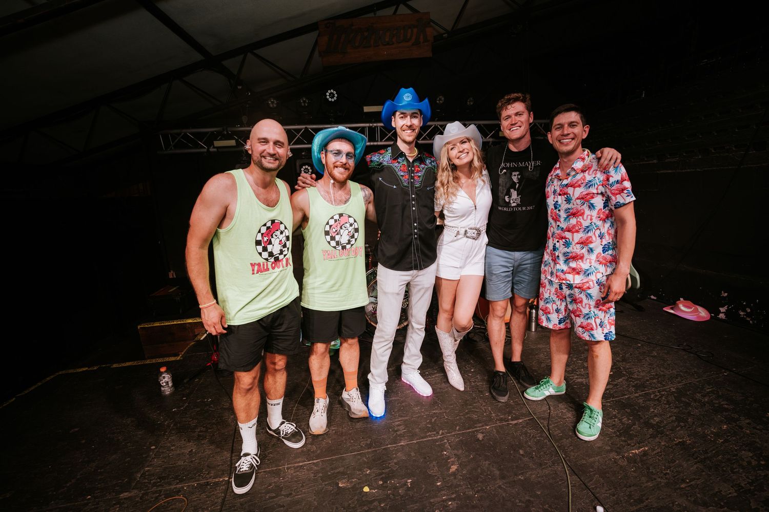 Group of friends in casual summer attire posing together on dark wooden stage.