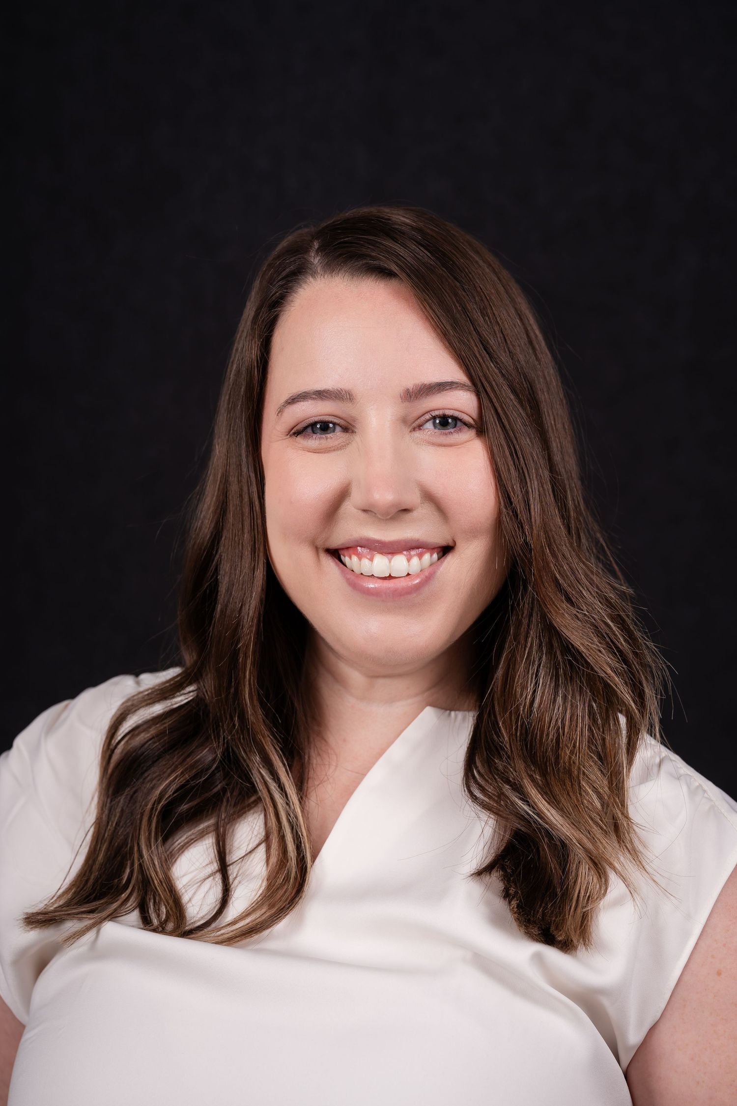 Professional headshot portrait of someone in a white blouse smiling warmly against a dark background.
