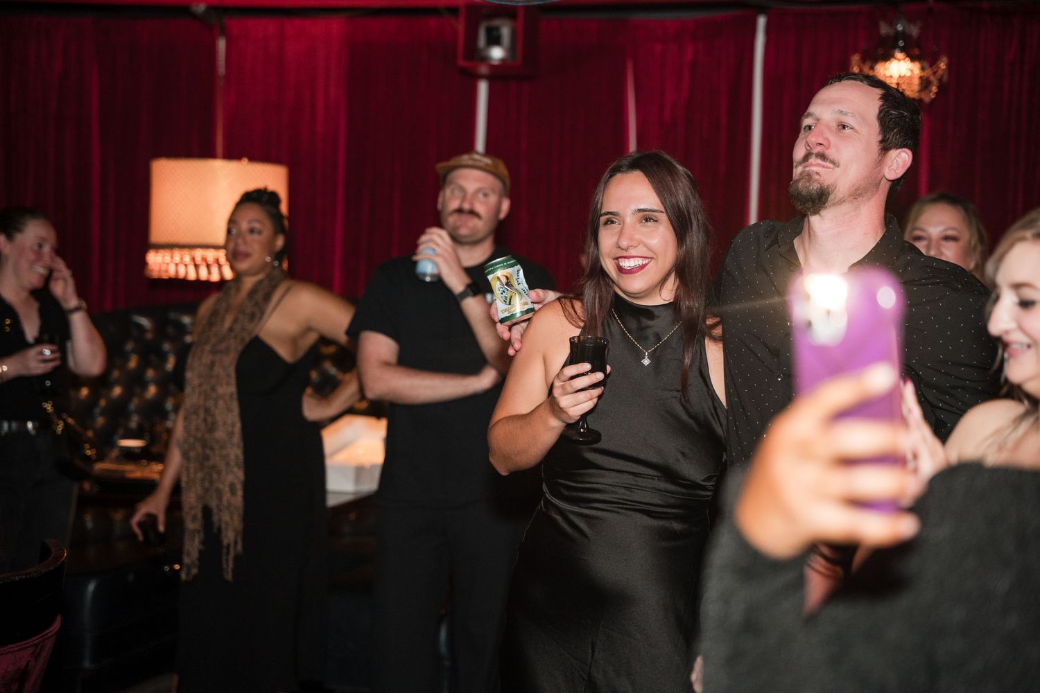Party guests laughing and taking photos with phones at a dimly lit celebration.