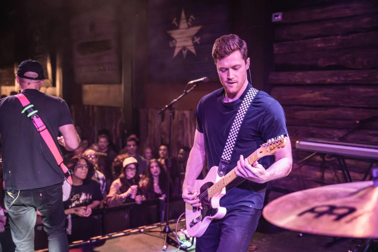 Musician playing electric guitar on wooden-walled stage during live performance.
