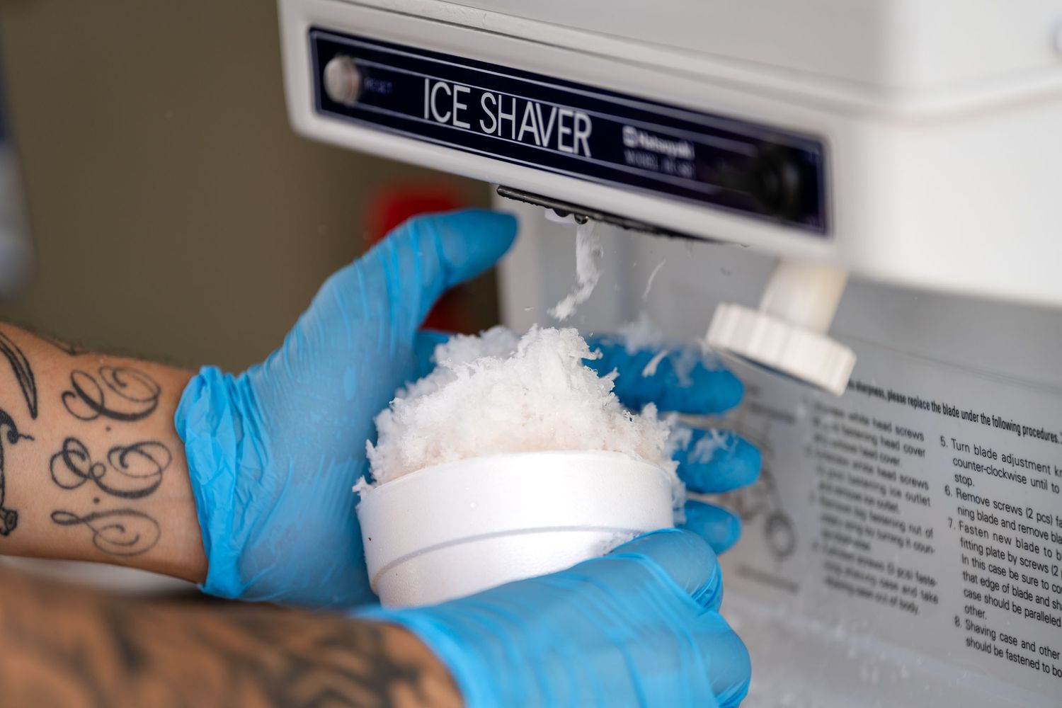 Hands wearing blue gloves scooping shaved ice from a commercial ice machine.