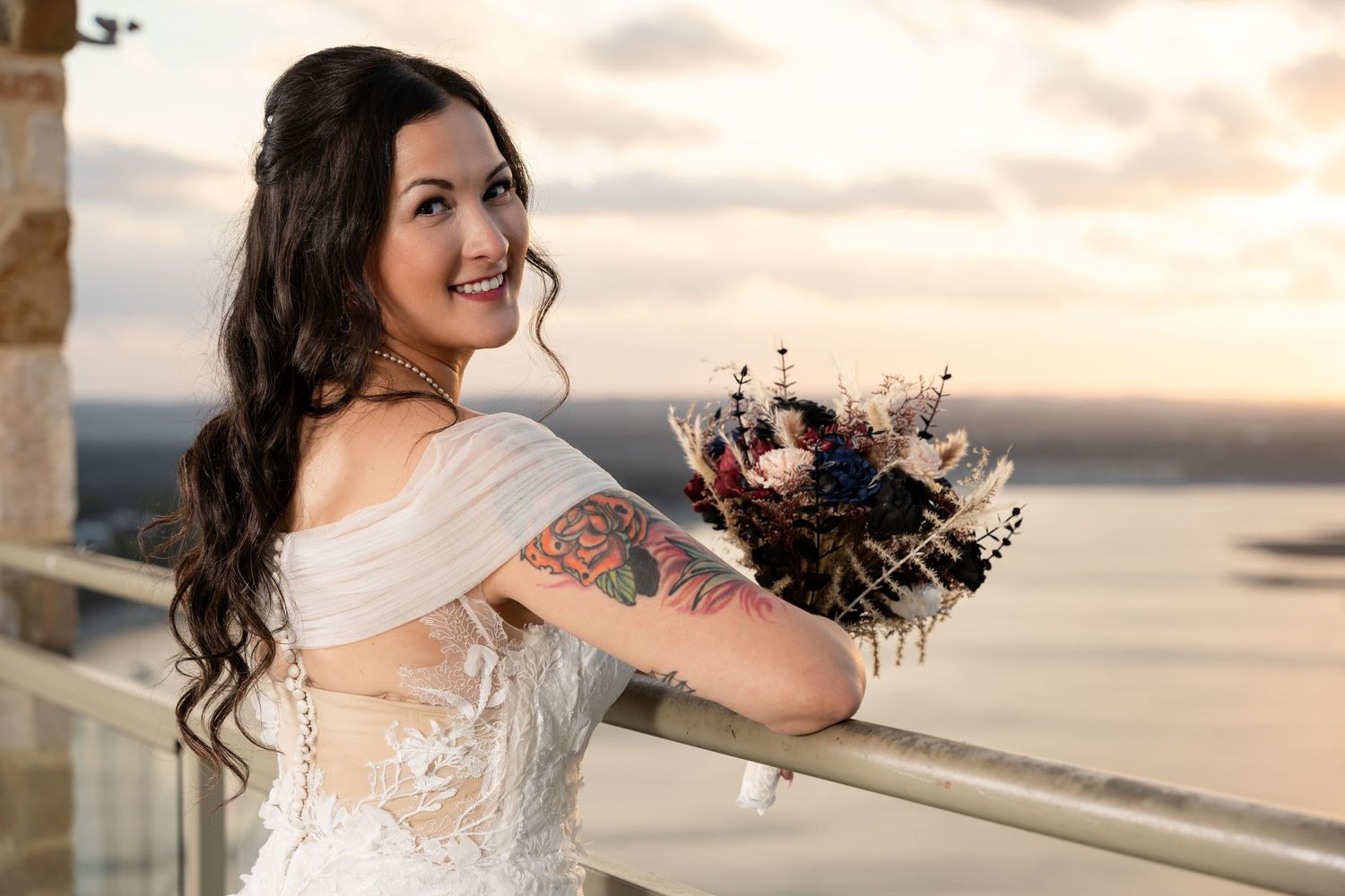 Bride in off-shoulder lace gown holding dark floral bouquet against sunset coastal backdrop.