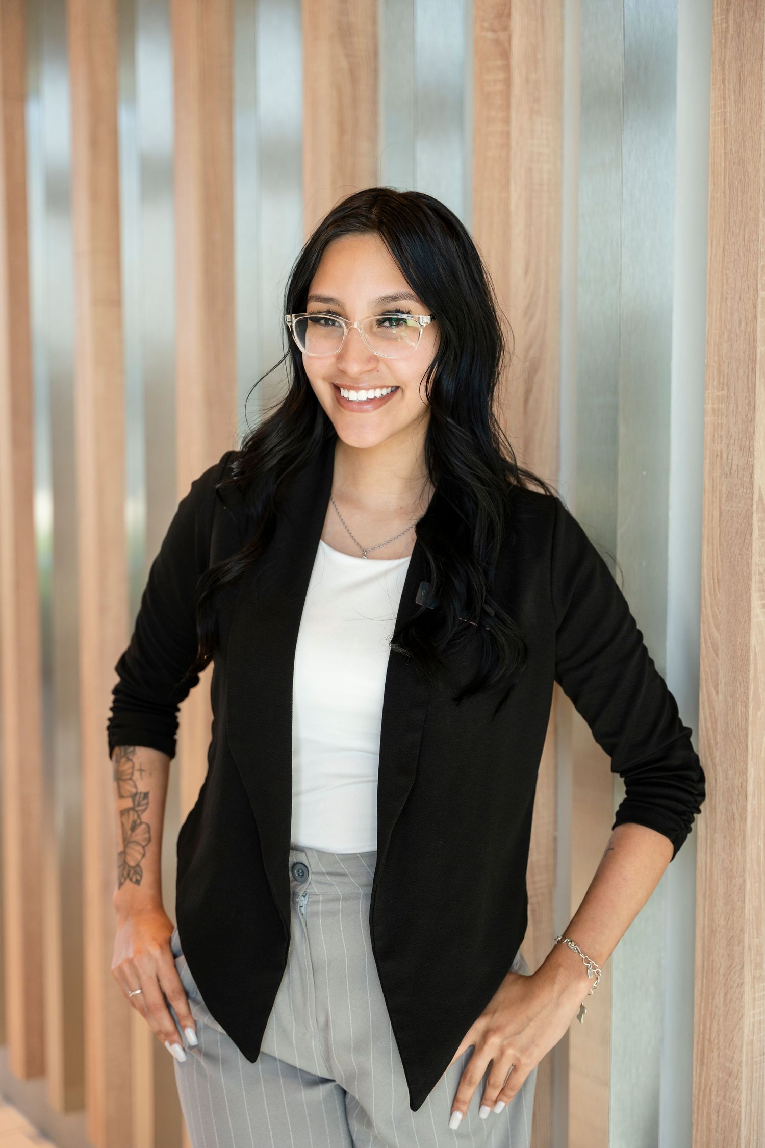 Professional portrait of someone in a black blazer, white top and gray pants standing by wooden slats with a warm smile.
