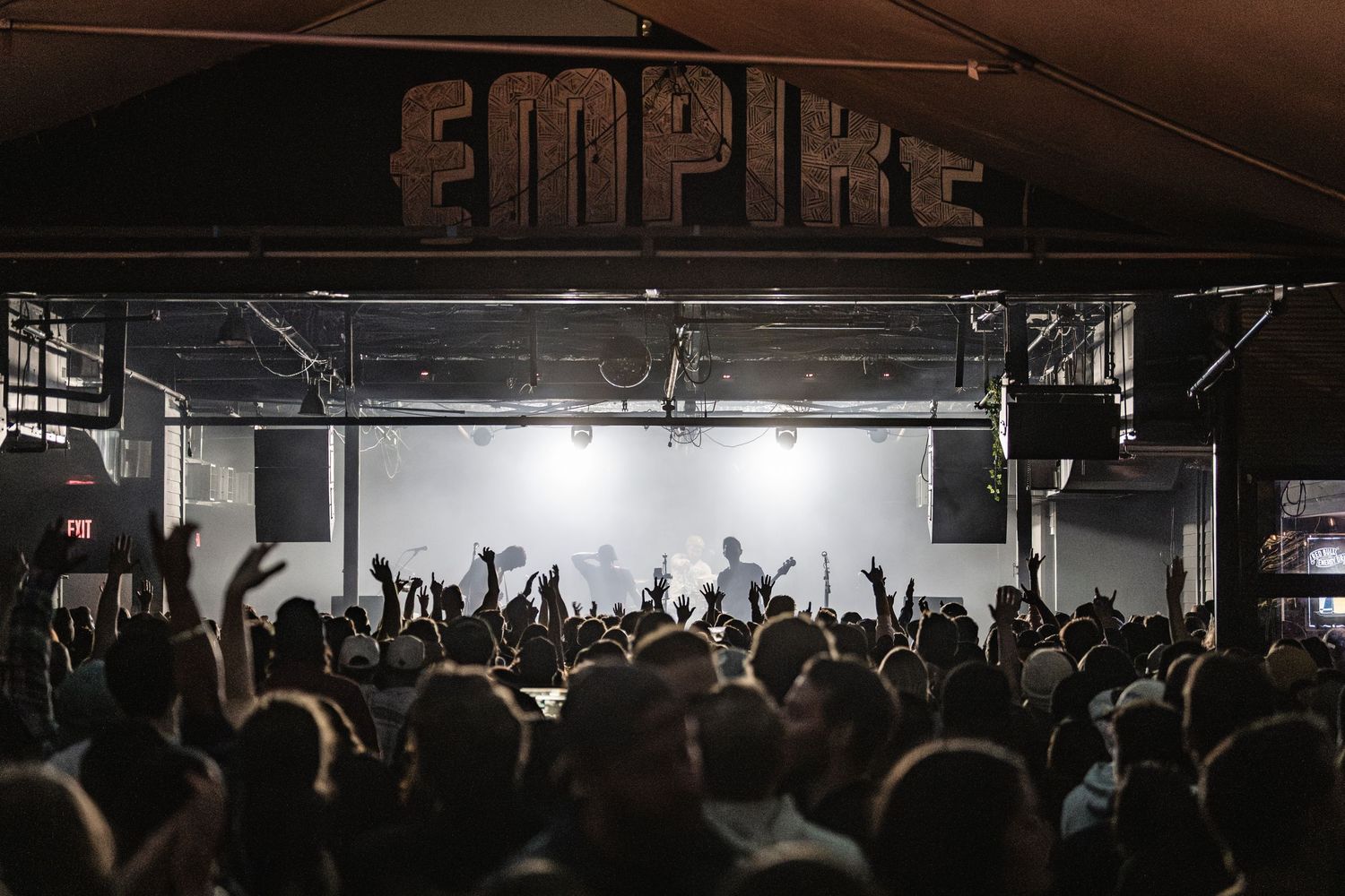 A crowded concert audience with raised hands silhouetted against bright stage lights under an Empire venue sign.