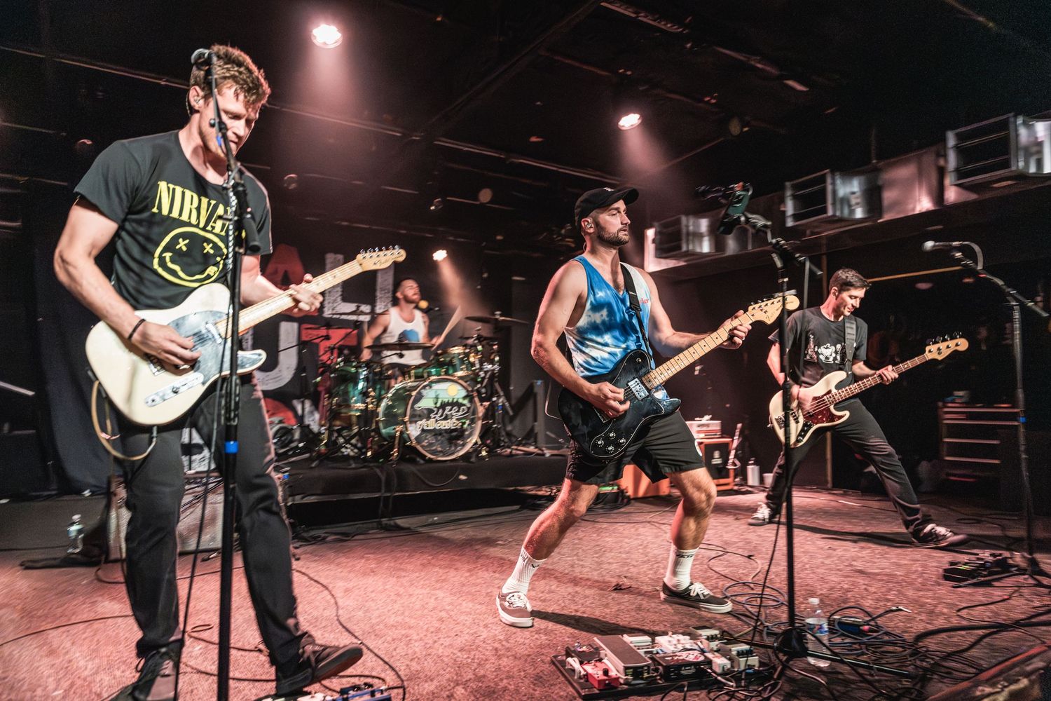 Rock band performing energetically on stage with guitars and drums under dramatic lighting.