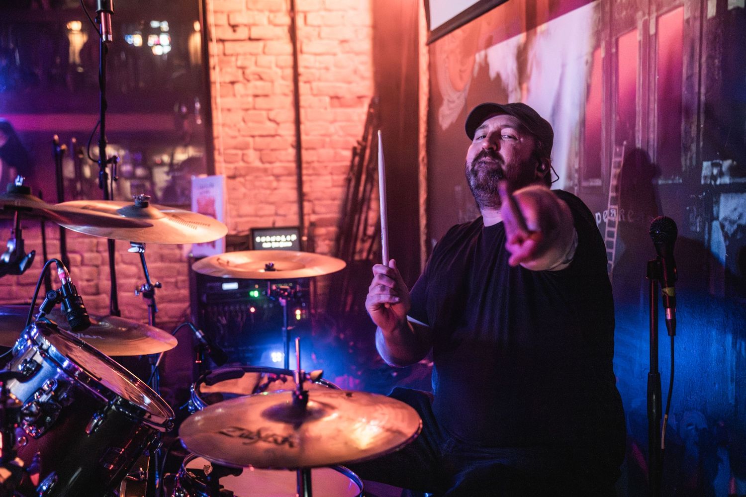 Drummer performs passionately on stage with dramatic lighting against brick wall backdrop.