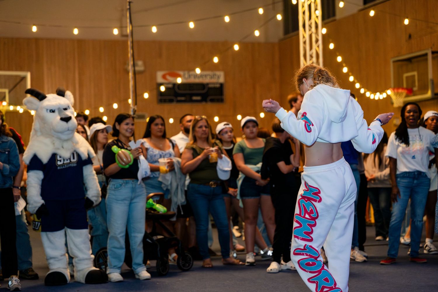 Mascots entertain a crowd of spectators at an indoor event with string lights illuminating the space.
