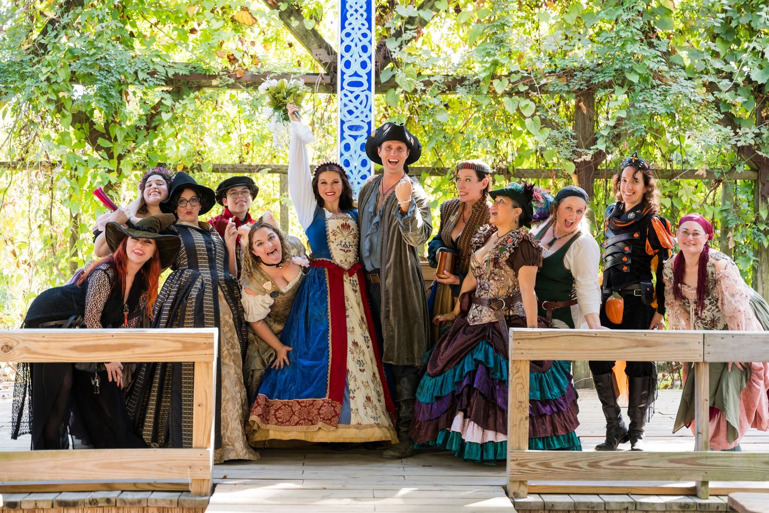 Group photo of renaissance fair performers in period costumes posing on wooden stage.