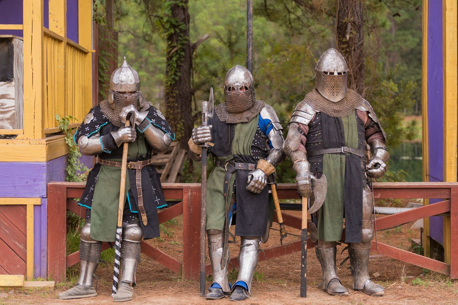 Three armored knights in medieval garb stand together outside a colorful building at a renaissance festival.