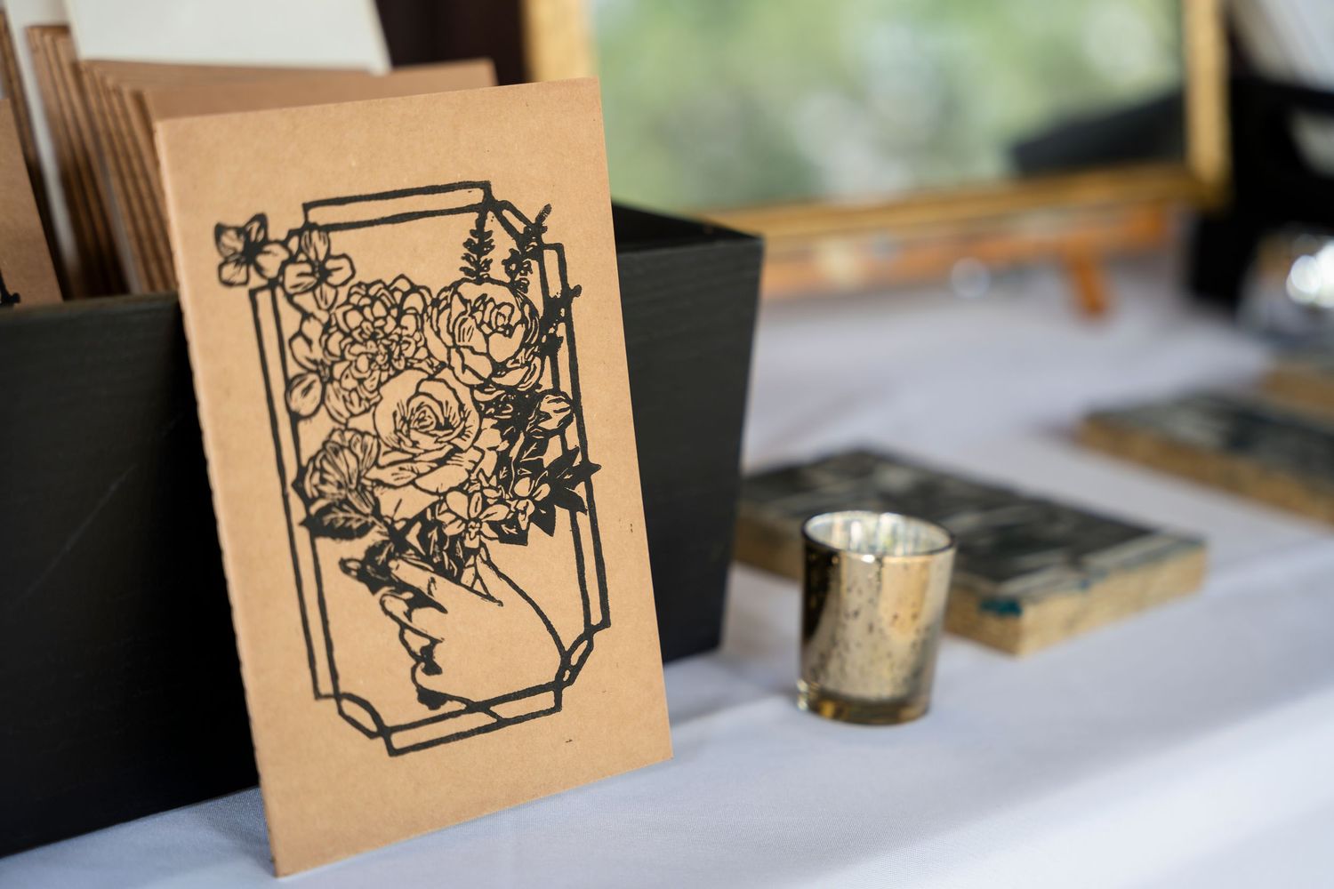 A kraft paper card with black floral illustration sits beside a small metallic candle holder on a white surface.
