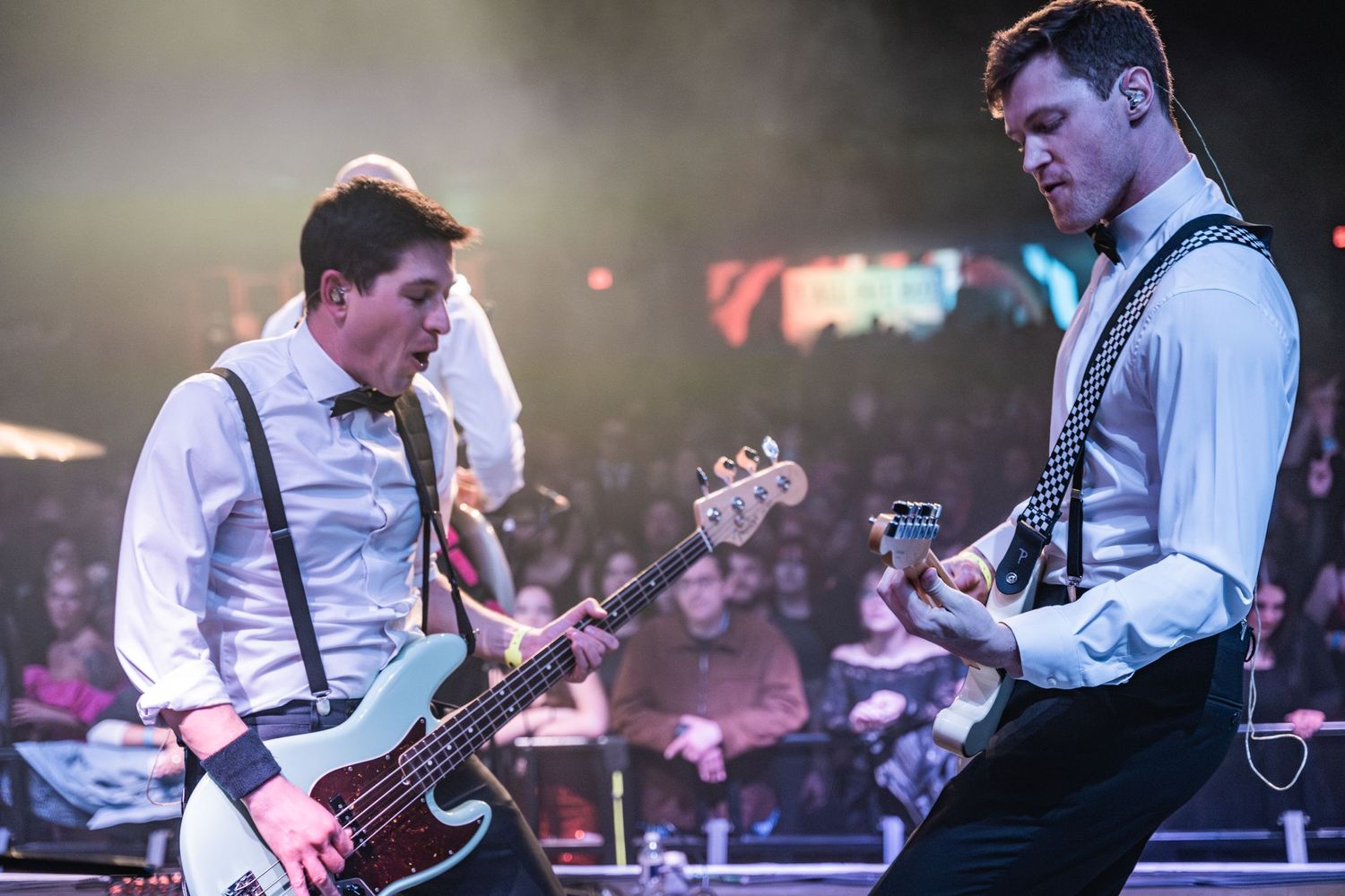 Two guitarists in vests and suspenders perform together on illuminated concert stage.