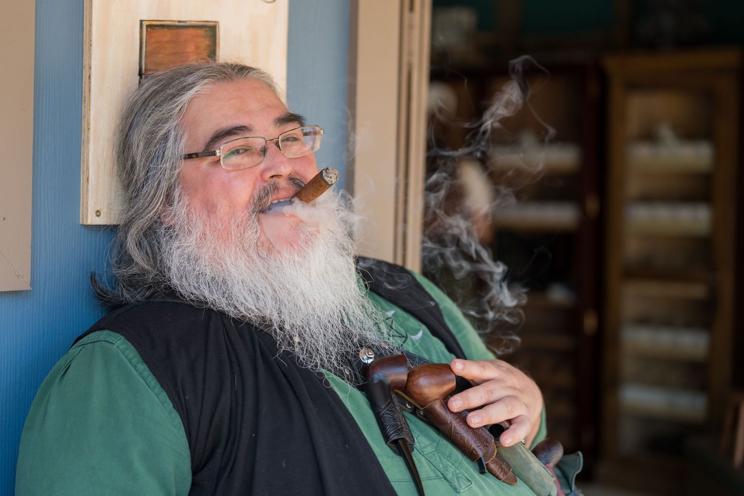 A gentleman with a long grey beard smokes a pipe while wearing medieval-style clothing.