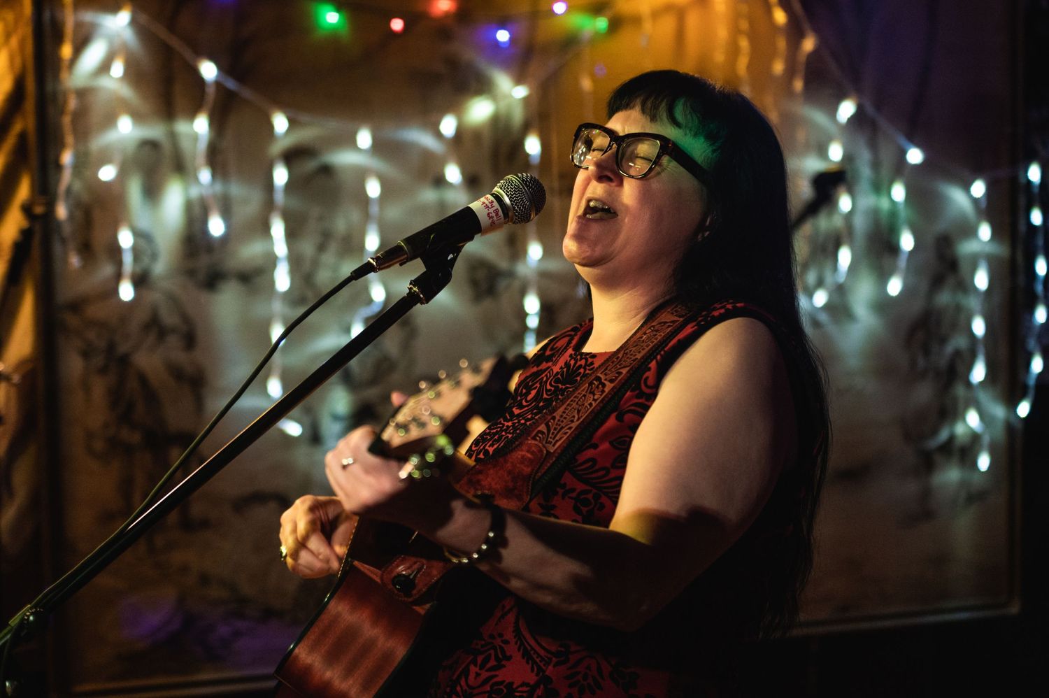 A musician performs with a guitar under colorful string lights in a dimly lit venue.