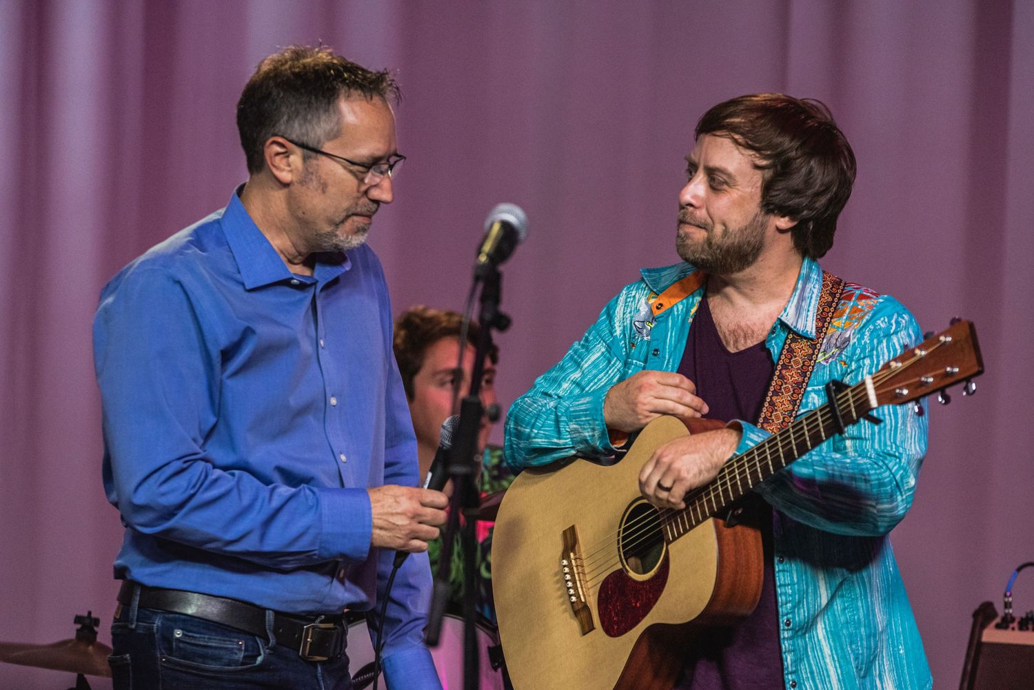 Two musicians perform on stage, one in blue shirt and one in turquoise playing acoustic guitar.