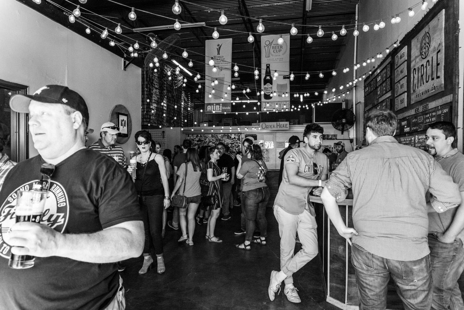 Black and white photo of crowd mingling under string lights in indoor venue.
