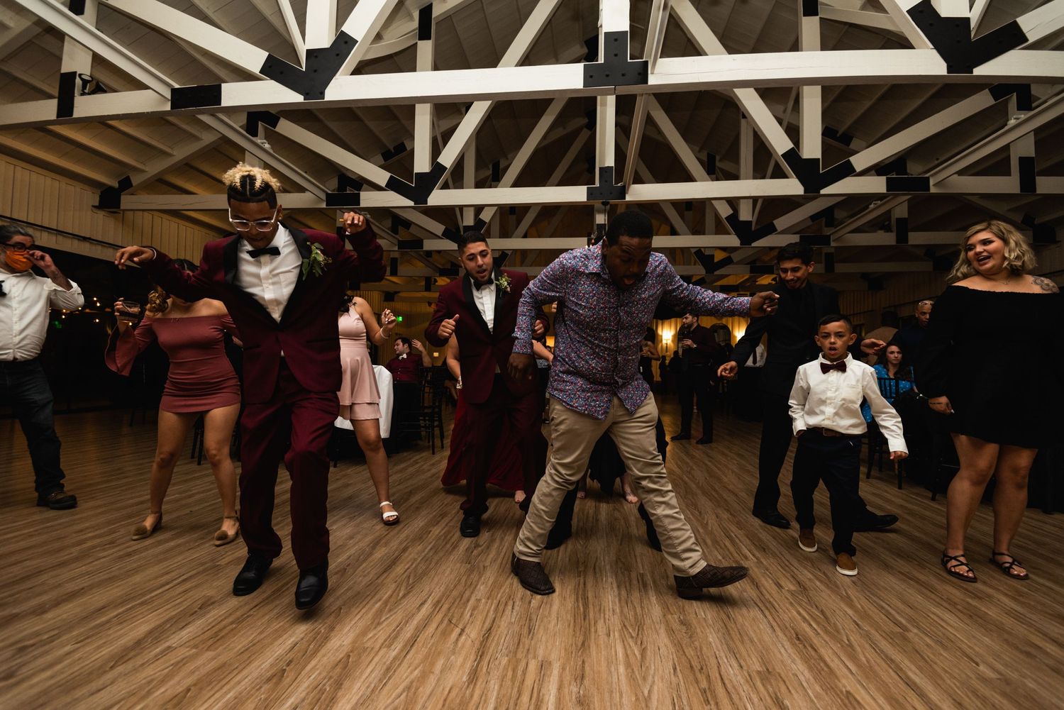 Party-goers dancing in formal attire on a hardwood floor under exposed wooden beams.
