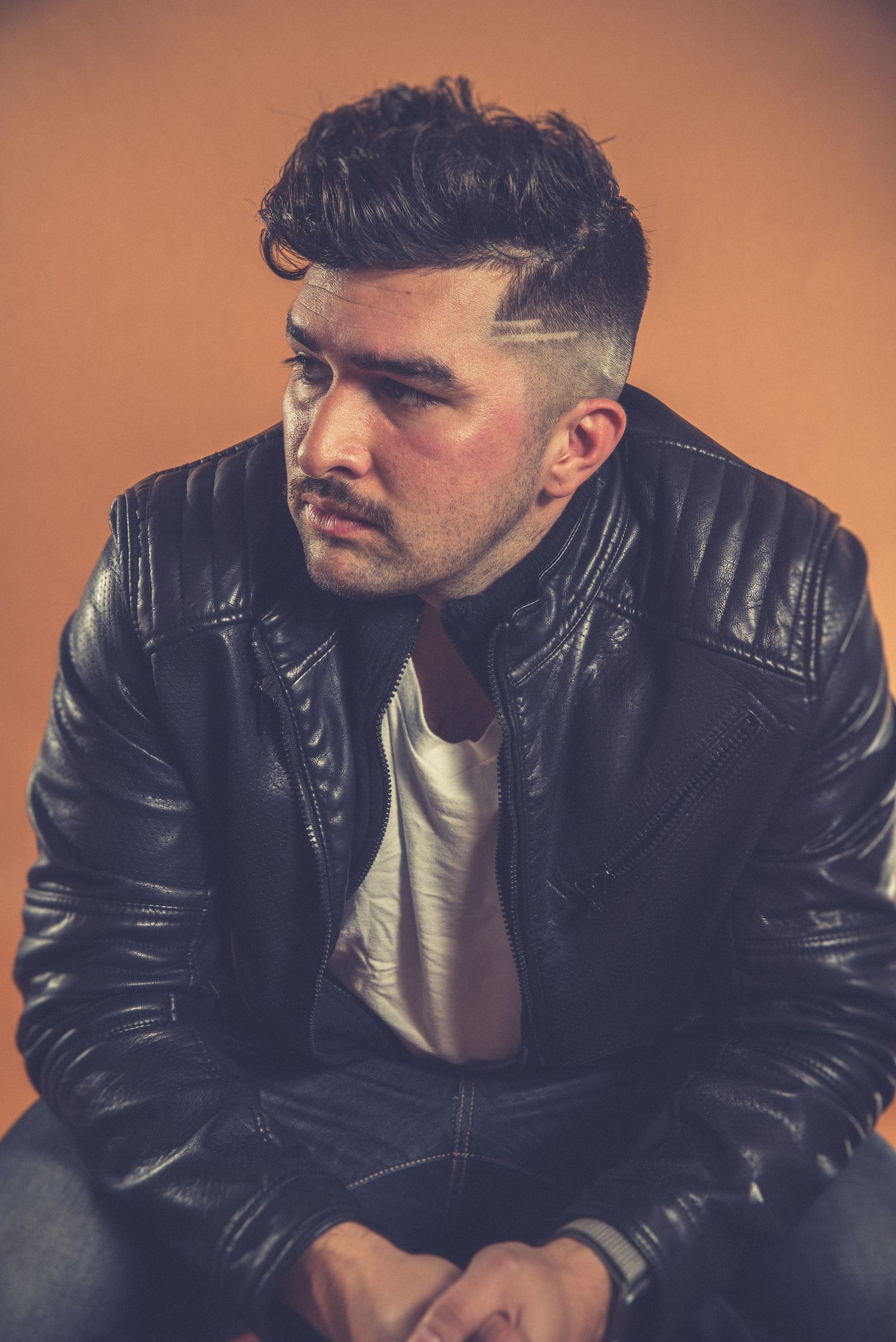 Portrait of someone in black leather jacket against orange background with contemplative expression.