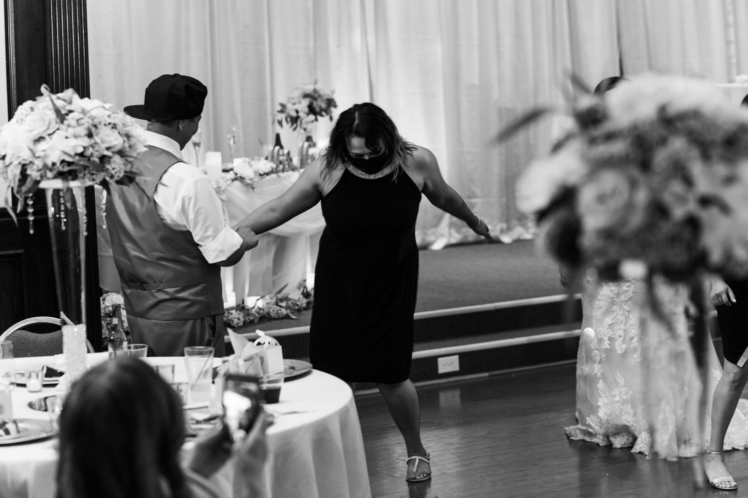 Black and white photo of guests dancing at a wedding reception with floral centerpieces visible.