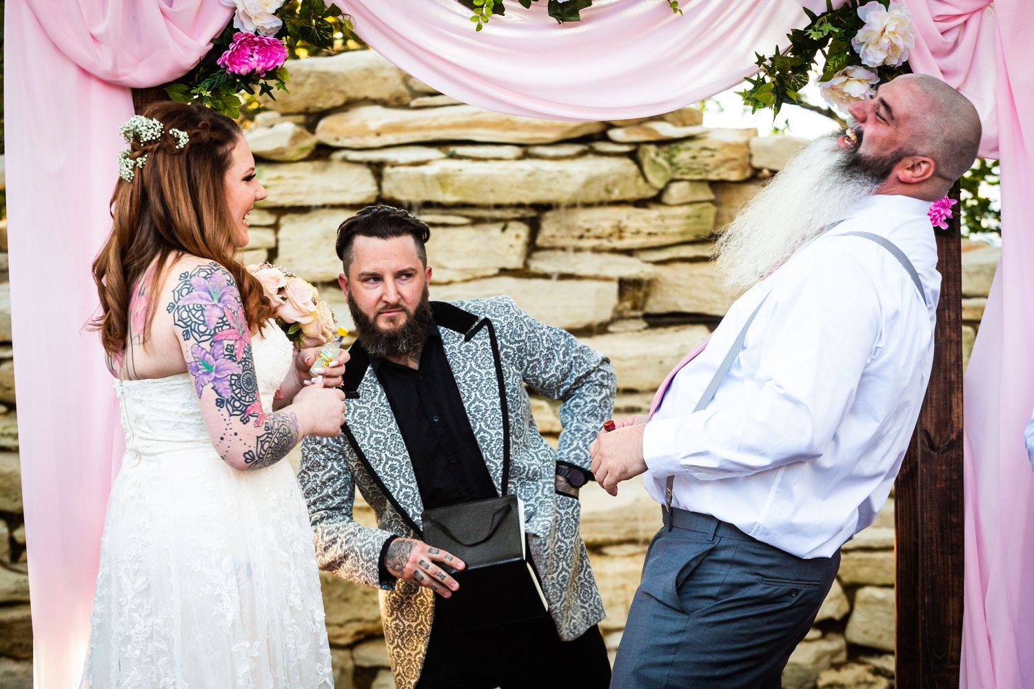 Wedding ceremony with officiant and couple in front of stone wall with pink draping and floral decor.