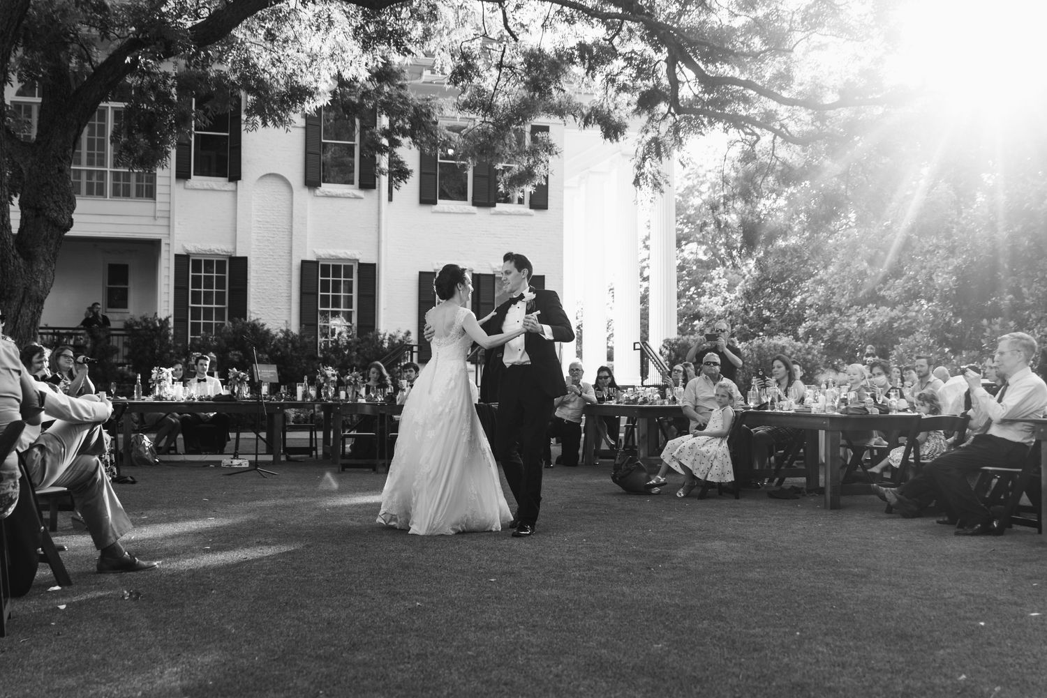 Black and white photo of a wedding reception dance on a lawn with a stately white house in the background.