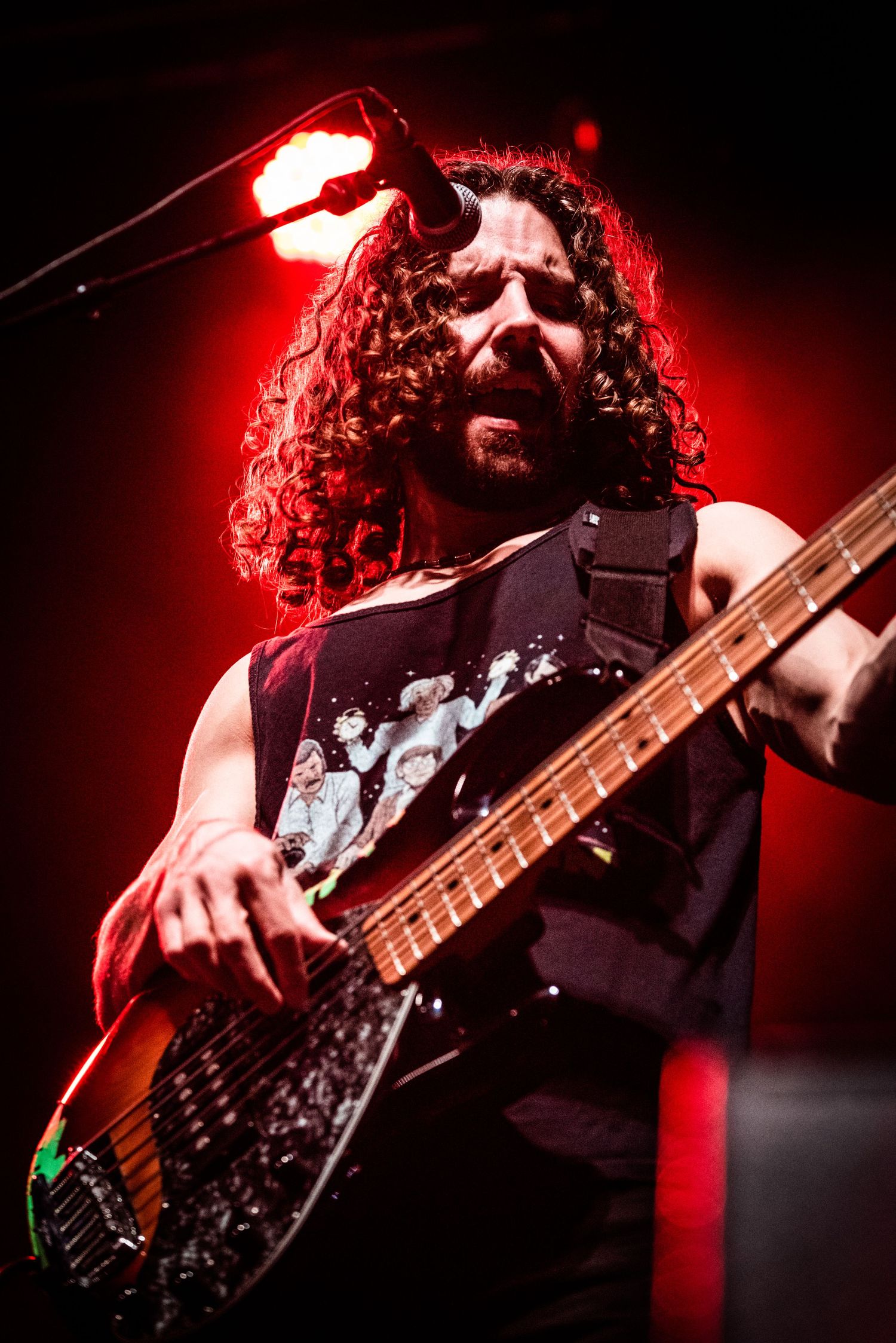 Bass player with long curly hair performs under red stage lighting.