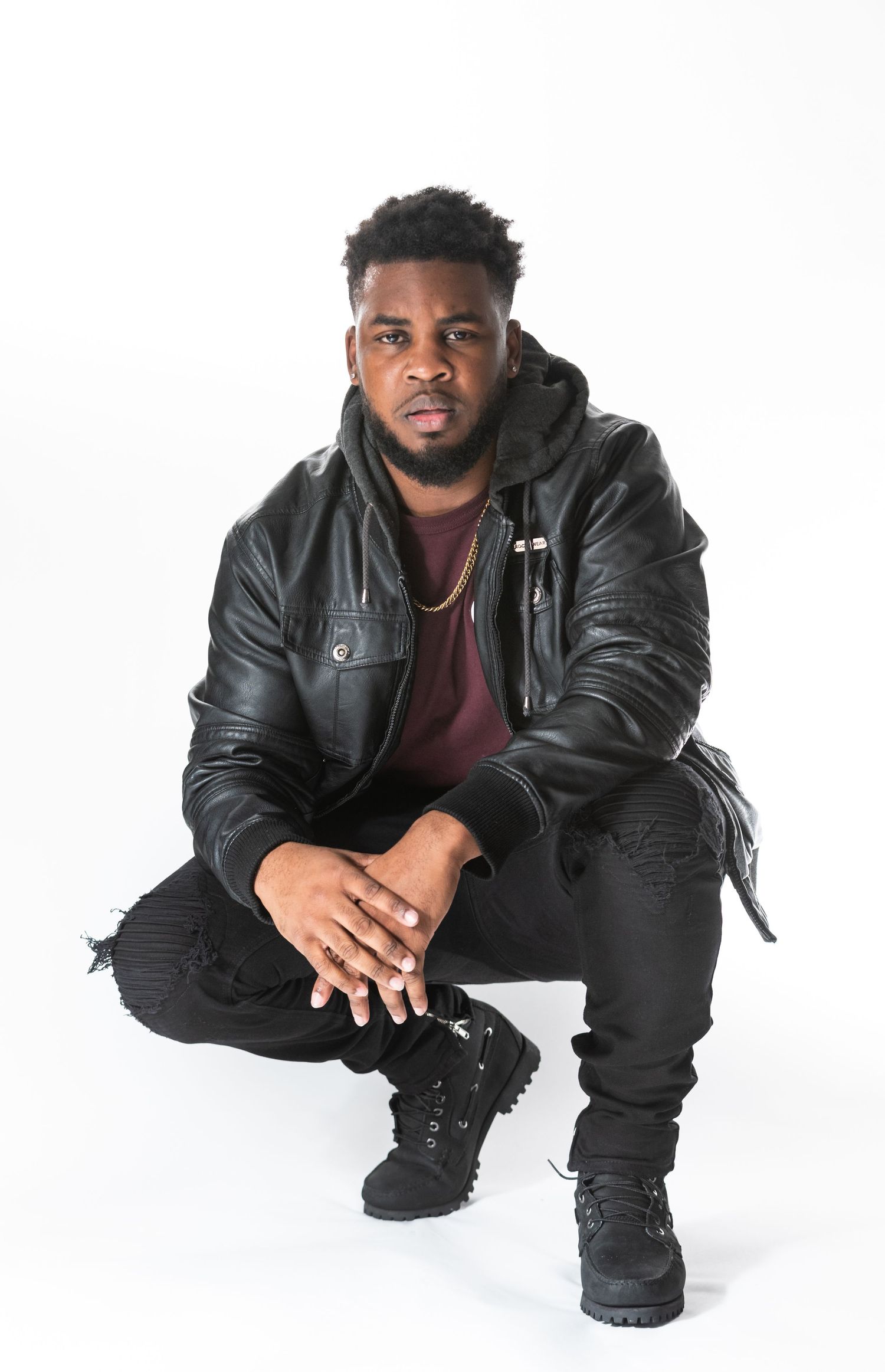 Urban style portrait featuring black leather jacket and ripped jeans in crouching pose against white background.
