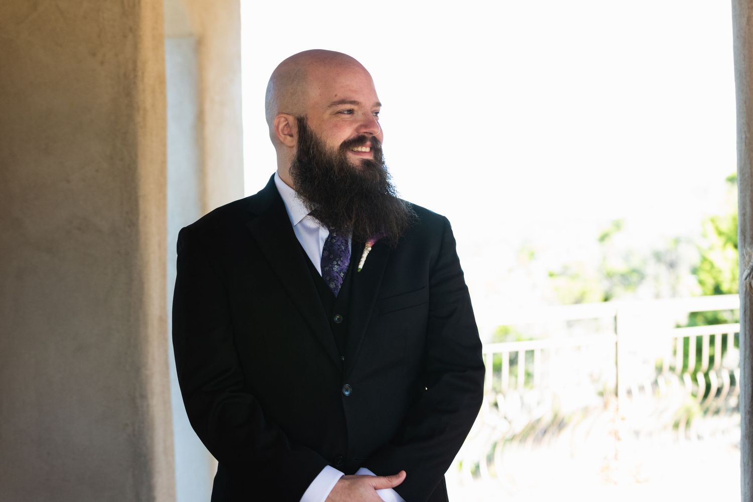 Man in black suit with full beard smiling during outdoor wedding ceremony.
