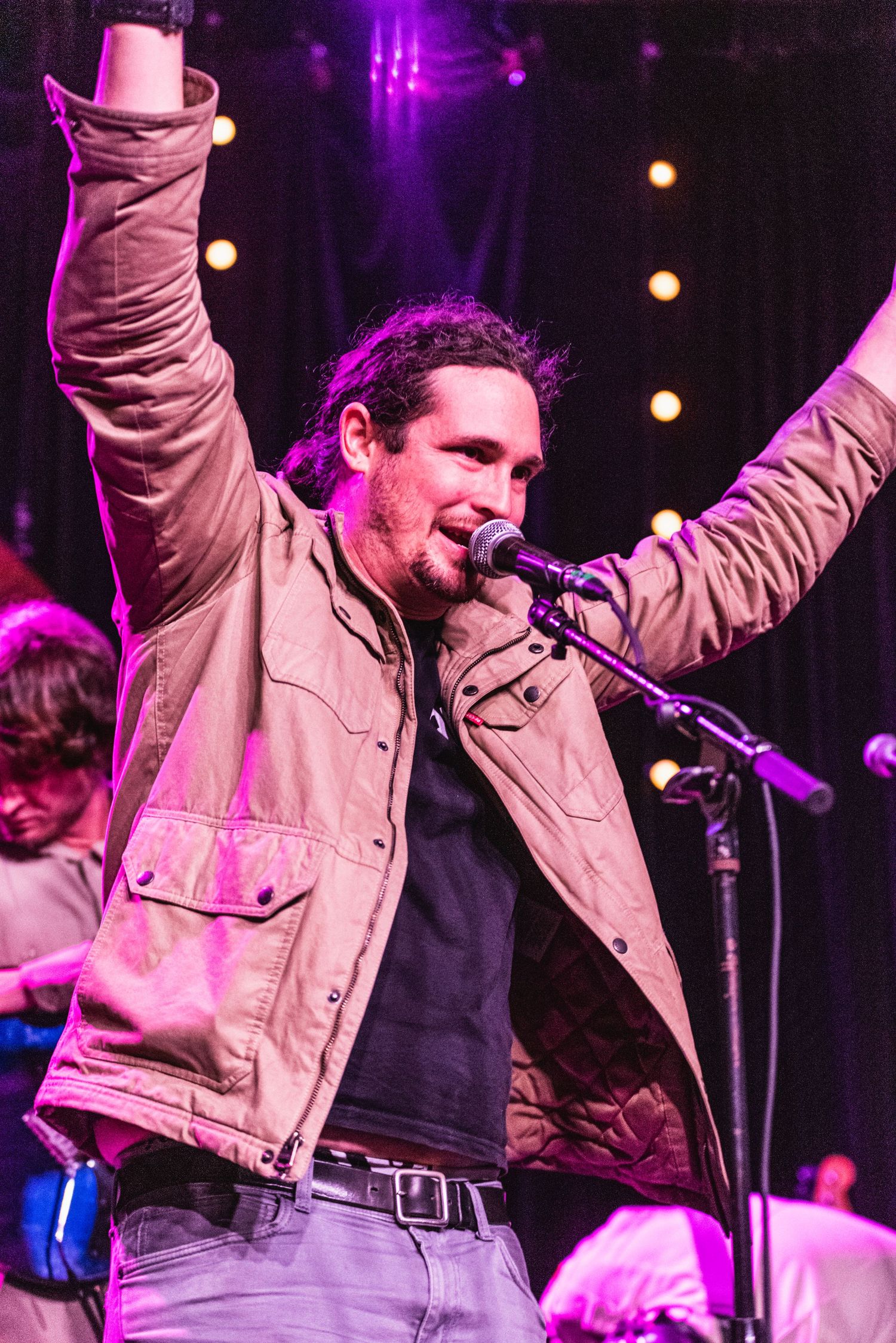 Energetic performer in tan jacket raises arms during concert performance under purple stage lights.