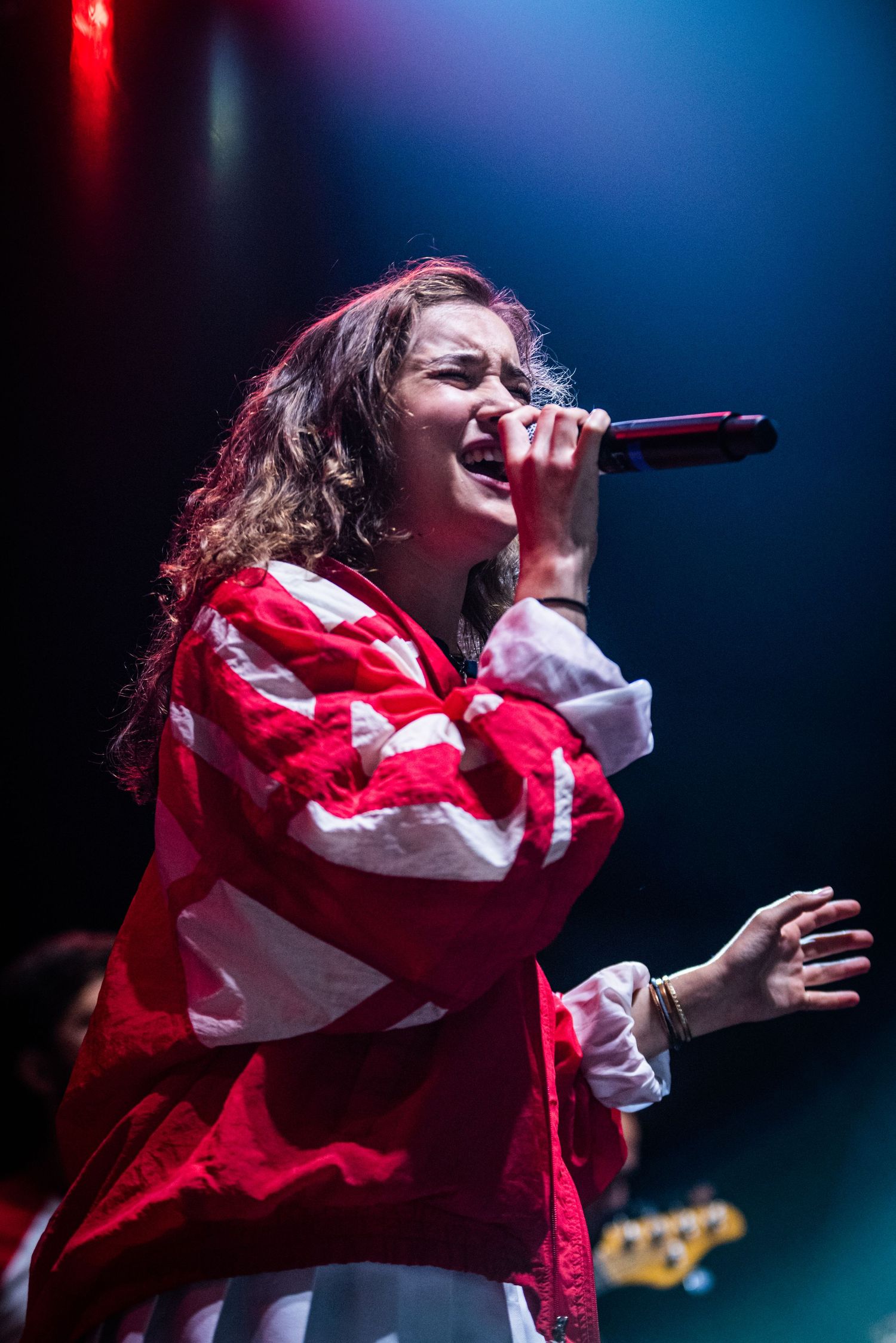 Performer wearing red and white jacket sings passionately into microphone on stage with blue lighting.