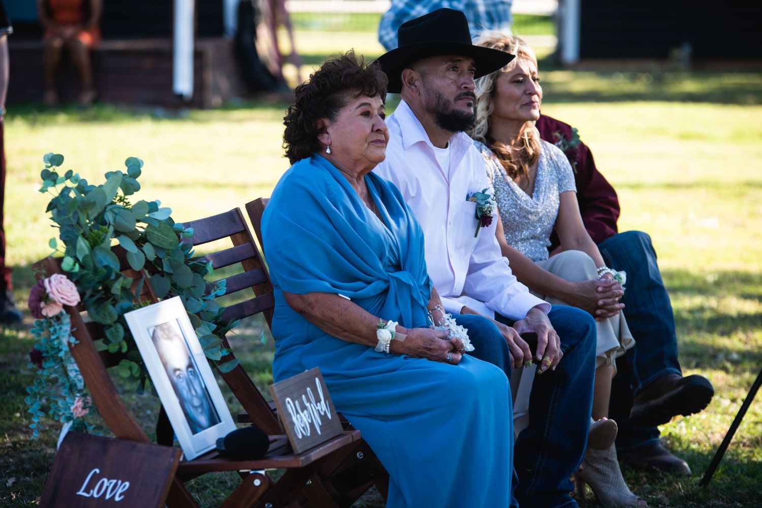 Wedding guests sit on wooden benches during an outdoor ceremony with framed photos displayed.
